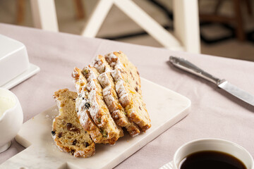 Traditional german christmas pastry stollen on a marble cutting board, coffee mug, butter plate on the table cloth and scandy style kitchen chair in the back
