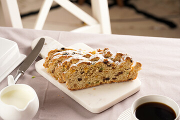Traditional german christmas pastry christstollen on a marble cutting board, coffee mug, butter plate on the table cloth and scandy style kitchen chair in the back