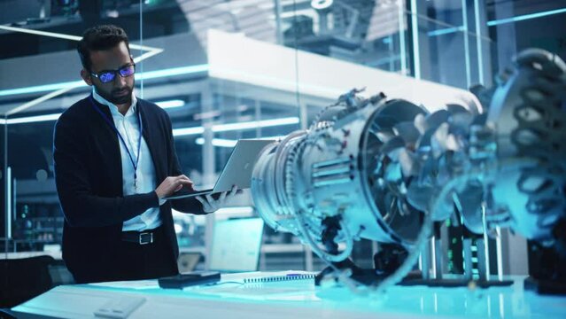 Young Industrial Engineer Working on a Futuristic Jet Engine, Standing with Laptop Computer in Scientific Technology Lab. Scientist Developing a New Electric Motor in a Research Facility.