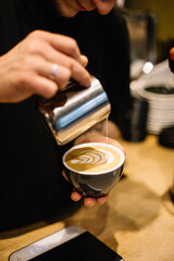Young male barista pouring frothed milk into the cup with freshly brewed double espresso coffee, making latte art, close up vertical view