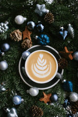 Delicious freshly brewed cappuccino coffee with a beautiful latte art on it, standing among silver, purple and blue ornamental balls, coconut stars, pine cones and green fir branches 