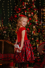 Pretty young girl in red winter dress and bow holds white present in the room with Christmas decoration. View from the back. Holiday concept