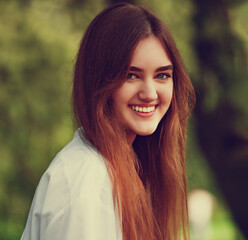 Beautiful toothy smiling teen woman looking happy outdoors in white shirt on summer green trees background. Closeup portrait in bright sunny day. Toned