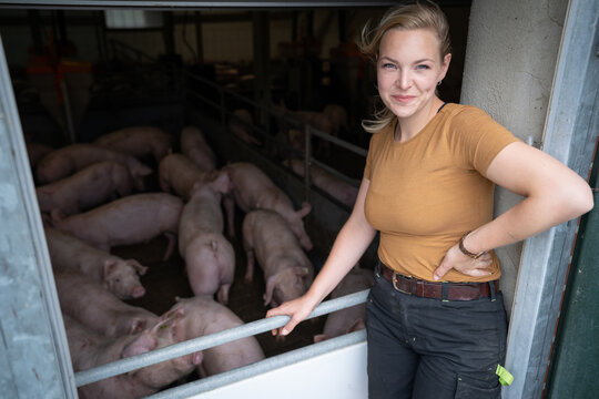 Junge hübsche Landwirtin steht vor einem offenen Schweinestall und freut sich über die gesunden Schweine.