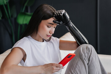 Tired young pretty woman leans on her bionic hand prosthesis eyes closed, sits on sofa. Upset girl...