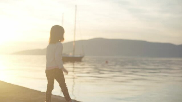A girl on a pier by the sea throws stones into the water against the backdrop of sunset