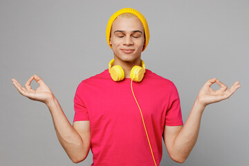 Young man of African American ethnicity he wear pink t-shirt yellow hat headphones hold spreading hands in yoga om aum gesture relax meditate try to calm down isolated on plain grey background studio.