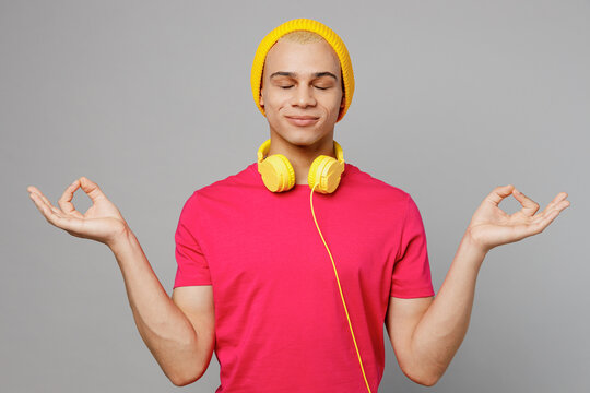 Young man of African American ethnicity he wear pink t-shirt yellow hat headphones hold spreading hands in yoga om aum gesture relax meditate try to calm down isolated on plain grey background studio. - Powered by Adobe