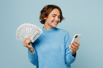 Young woman wear knitted sweater use mobile cell phone hold in hand fan of cash money in dollar...