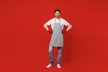 Full body smiling happy fun confident cool young male housewife housekeeper chef cook baker man wear grey apron oven mittens stand akimbo solated on plain red background studio. Cooking food concept.