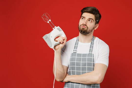 Young Male Housewife Housekeeper Chef Cook Baker Man Wear Grey Apron Hold Mixer Going Make Cream Or Beat Egg Whites Look Overhead On Area Isolated On Plain Red Background Studio. Cooking Food Concept.