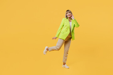 Fototapeta na wymiar Full body feminine elderly woman 50s years old wearing green jacket white t-shirt stand on one leg hold face look camera isolated on plain yellow background studio portrait. People lifestyle concept.