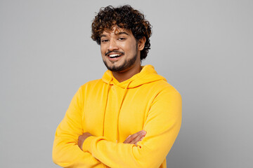 Young confident smiling happy cheerful fun Indian man 20s he wear casual yellow hoody hold hands crossed folded look camera isolated on plain grey background studio portrait People lifestyle portrait