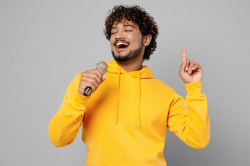 Young cheerful Indian man 20s he wearing casual yellow hoody sing song in microphone on karaoke...
