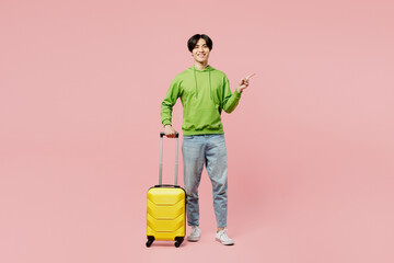 Full body traveler man of Asian ethnicity wear green hoody hold suitcase valise isolated on plain pastel pink background Tourist travel abroad in free spare time rest. Air flight trip journey concept