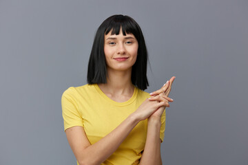 a cute, playful woman poses on a gray background standing in a yellow cotton T-shirt and gesturing with her hands in a relaxed manner, smiling pleasantly