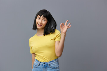 a funny, sweet woman with short black hair stands on a gray background in a yellow T-shirt and shows the OK sign with her hands, smiling at the camera. Horizontal photo with empty space