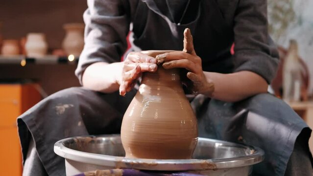 Close-up of potter's hands covered with clay making beautiful vase on throwing wheel in pottery workshop. Creativity and traditional crafts concept. Handmade. Clay