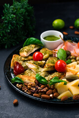 Set to wine cheese bread with pesto, pork, assorted cheeses, capers, olives, tomatoes, pesto sauce, walnuts, almonds, basil. Vertical orientation, closeup.