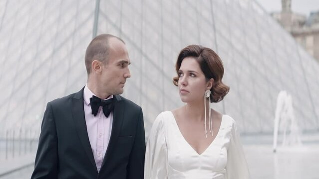 Beautiful elegant couple of lovers walking in ancient city. Action. Luxurious couple walks in love in Paris. Couple in elegant outfits walks on background of glass pyramid