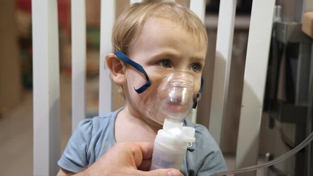 Sick Baby Girl with making Inhalation with a Mask on his Face sitting in Living Room at Home. Children Healing. Child Inhalation therapy with Compressor nebulizer. Inhaling fumes medication into lungs
