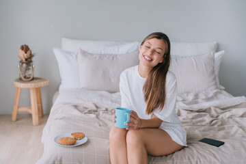 Obraz na płótnie Canvas Adorable caucasian young woman sitting on bed holds cup of coffee eyes closed smiles. Close up of beautiful Italian girl enjoys sunny morning at home. Happy people. Domestic leisure. Cozy home.