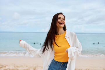 Portrait of a happy woman smile with teeth with long hair brunette walks along the beach in a yellow tank top denim shorts and a white shirt by the sea summer travel and feeling of freedom, balance