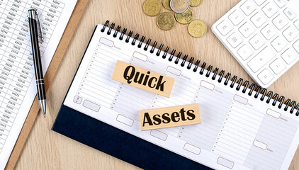 QUICK ASSETS word written on wooden block on planner with coins, clipboard and a calculator