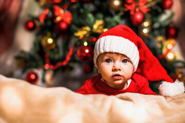 Joyful baby looking at camera in Christmas photo. Newborn baby in red cap of Santa Claus and Christmas tree in background