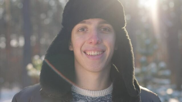 Headshot portrait of positive Caucasian young man in earflaps hat standing in sunshine looking at camera smiling. Cheerful millennial guy posing in slow motion in winter forest in sunbeam
