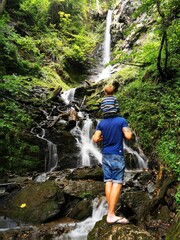 Wandernde r Mann steht an einem Wasserfall mit Kind auf den Schultern 