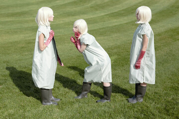 Conceptual photo of identical blond girls in clothing covers, pink gloves and rubber boots standing on green field and looking in the mirror 