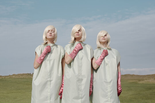 Conceptual portrait of identical blond girls wearing clothing covers and pink gloves standing outdoors in same positions with their eyes closed 