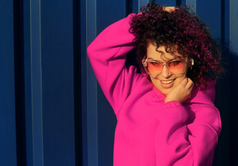 Fototapeta na wymiar Happy young woman with curly hair in magenta suit and sunglasses posing on blue background.