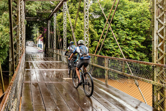 Ciclistas atravessando ponte de madeira sobre rio em dia de chuva