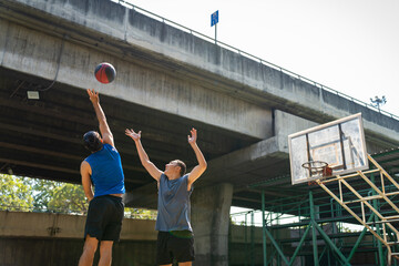 Fototapeta na wymiar Two man athlete playing streetball match shooting and defense basketball on outdoors court together in sunny day. Sportsman do sport training basketball at street court under highway in the city.