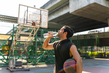 Caucasian sportsman basketball player drinking and pouring water from a bottle on his face after do sport training and playing streetball on outdoors court under highway in the city in sunny day.