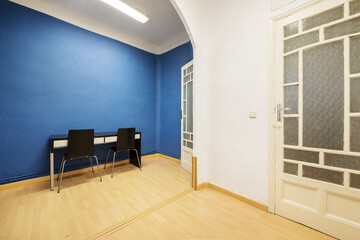 A room with old solid wood doors with translucent windows, blue and white walls and a black desk with white drawers, a plaster arch and laminated flooring
