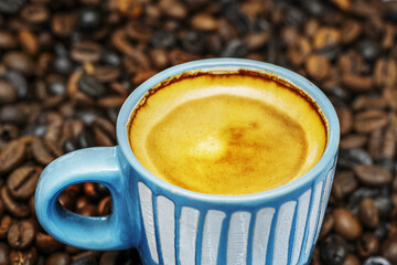 Close-up of a blue cup of espresso coffee with froth on top of a pile of roasted coffee beans