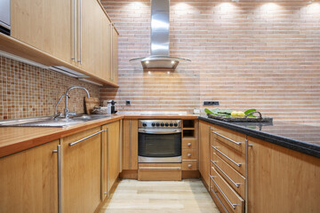 Kitchen with wooden cabinets arranged in a u-shape with a wooden and black marble countertop and a stainless steel extractor hood on an exposed brick wall