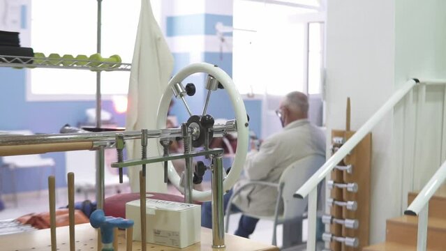 Focusing on various diagnostic and treatment equipment in the foreground inside of a physiotherapy clinic as the doctor is observing a patient in the background, cinematic slow motion side tracking