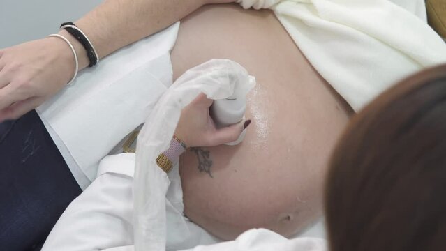 Top view of a female ultrasound technician doing a live sonogram on a pregnant caucasian client lying down on a bed table, close up with no face