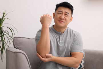 Asian man suffering from pain in his elbow on armchair indoors