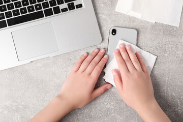 Woman wiping smartphone with paper towel at gray table, top view