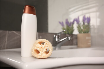 Natural loofah sponge and bottle of shower gel on washbasin in bathroom, space for text