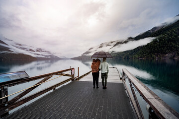 
A young sympathetic couple stands with an umbrella in their hands against the backdrop of a beautiful lake and mountains.