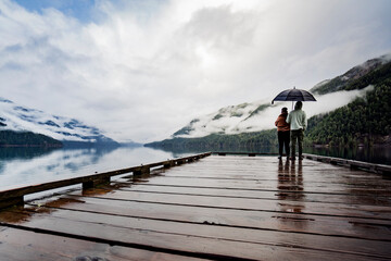
A young sympathetic couple stands with an umbrella in their hands against the backdrop of a beautiful lake and mountains.
