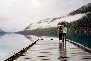 
A young sympathetic couple stands with an umbrella in their hands against the backdrop of a beautiful lake and mountains.