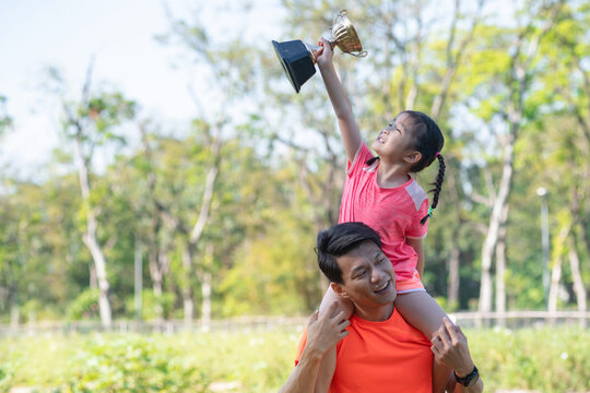Asian little girl sit on father shoulder was overjoyed with a trophy and lifted the trophy over her head, showing at outdoor park