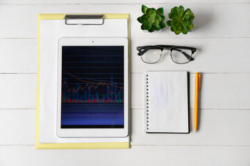 Tablet computer with stock data, clipboard, eyeglasses and houseplants on white wooden background. Finance trading